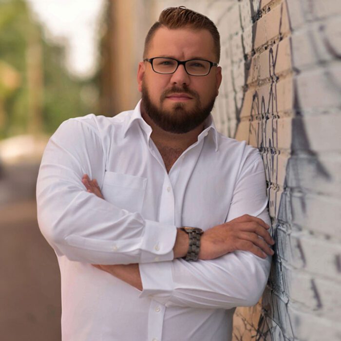 David Aaron leaning against a wall with graffiti wearing a white dress shirt crossing his arms